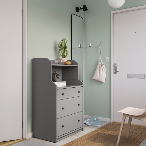 A grey HAUGA chest with 3 drawers and a top shelf against a green wall. It includes a mirror, plant, light, and a shoe rack.