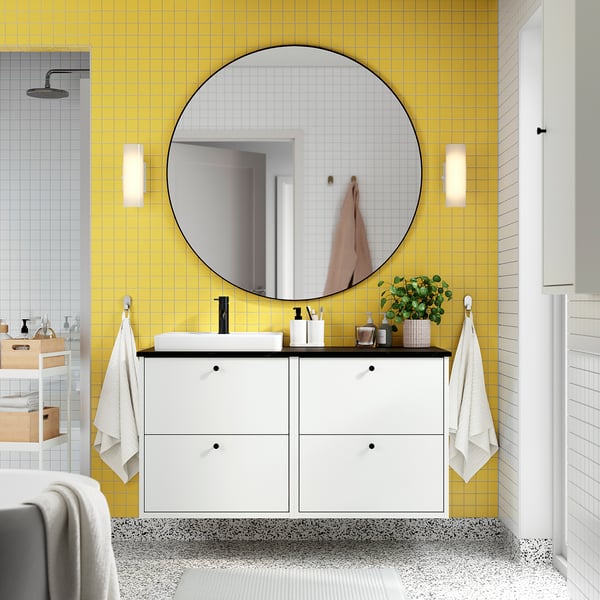 Modern bathroom with yellow tiled walls, large round mirror, white storage cabinet, rectangular sink, small plants, and wall-mounted towel hooks.