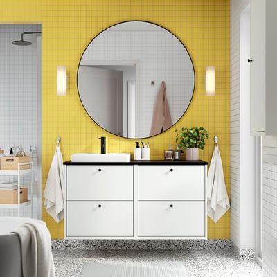 Modern bathroom with yellow tiled walls, large round mirror, white storage cabinet, rectangular sink, small plants, and wall-mounted towel hooks.