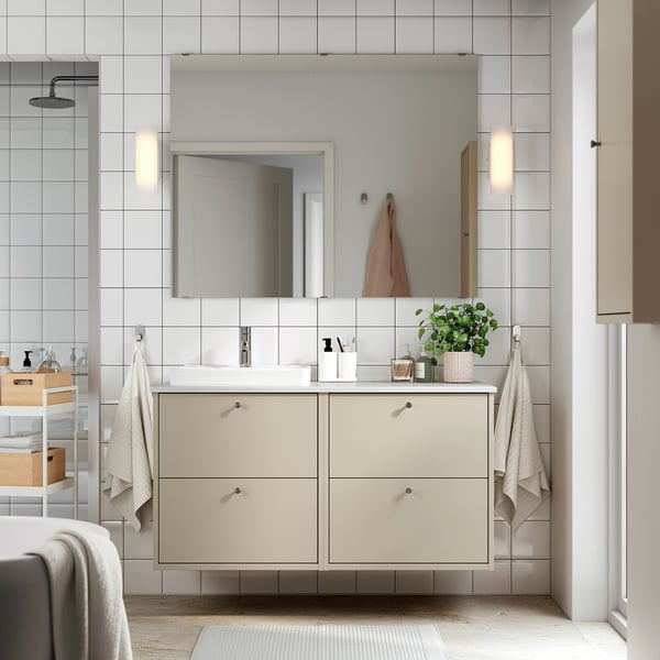 A modern beige bathroom vanity with white countertop, mirror, and wall lights. Dual basins and storage, with towels hanging beside.