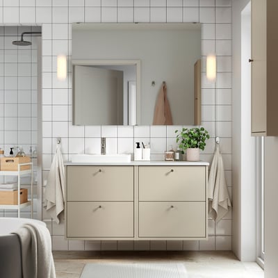 A modern beige bathroom vanity with white countertop, mirror, and wall lights. Dual basins and storage, with towels hanging beside.