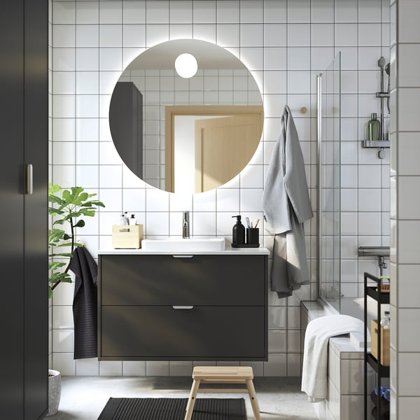 Modern bathroom: HAVBÄCK black unit, round mirror, wood stool, towels, plant, glass shower.