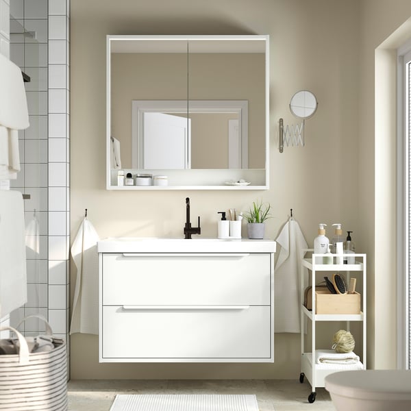 Modern white vanity with drawers and ceramic counter, large mirror. Black tap, rolling cart for storage.