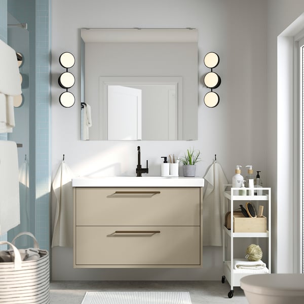 Beige HAVBÄCK vanity, white counter, black tap, large mirror, two drawers, wall lights, and side cart for toiletries.