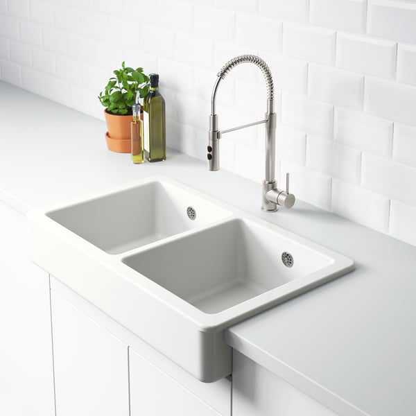 A modern kitchen sink with a white double basin and sleek tap, featuring a green plant and two glass bottles beside it.