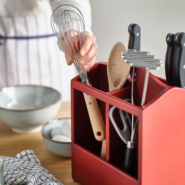 Person removing a whisk from a red wooden kitchen utensil holder. Bowl, small dishes, and towel are nearby.