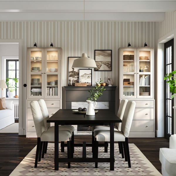 Cosy dining area with black HAVSTA table, beige chairs, grey cabinet, white pendant lamp, light-striped walls, and wooden floor with rug.