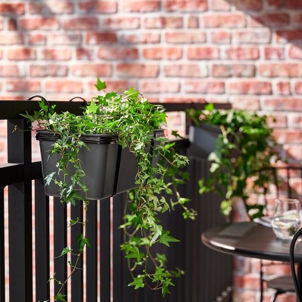 Black hanging plant pots on railings with lush greenery against a brick wall.