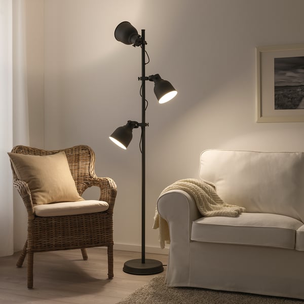 HEKTAR floor lamp, black, industrial design, large metal shades, adjustable height, stands tall beside wicker chair and beige sofa.