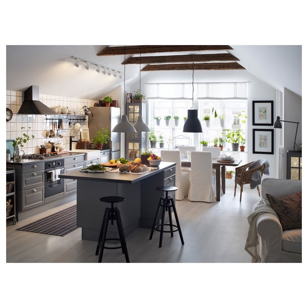 Modern kitchen with factory lamps over dining table, wooden beams, white walls, grey cabinets, cosy living area with sofa, wicker chair, and plants.