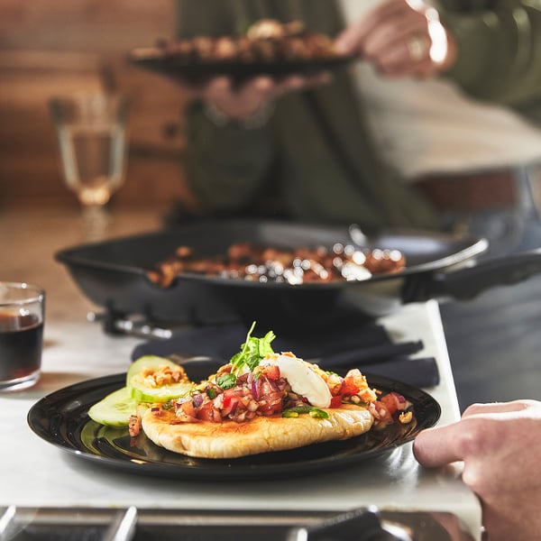 A person holding a plate of grilled food with avocado slices and bacon on a table with a grill pan and drinks.