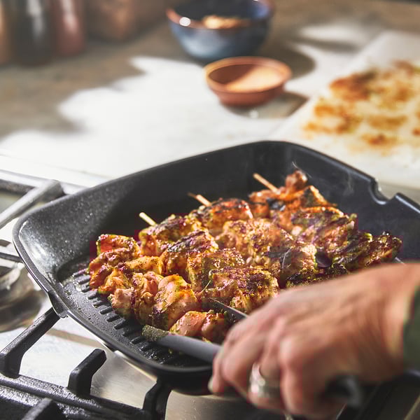 Person grilling meat skewers on a non-stick pan with grips, showcasing even heat distribution and easy handling.