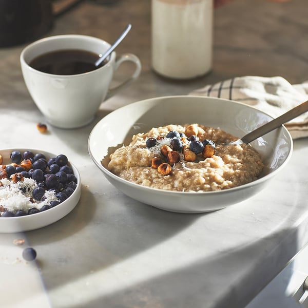 Bowl of oatmeal topped with blueberries and nuts, beside a cup of coffee and a smaller dish with blueberries, coconut flakes, and nuts.