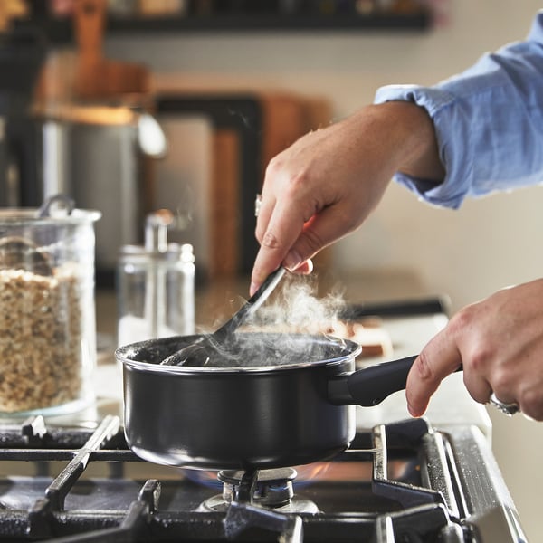 Person cooking with a HEMLAGAD saucepan on a stove. Steam rises as they stir the food with a spatula.