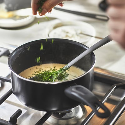 Image description: a hand adds chopped herbs into a saucepan on a stove, showcasing its use for cooking sauces and heating food.