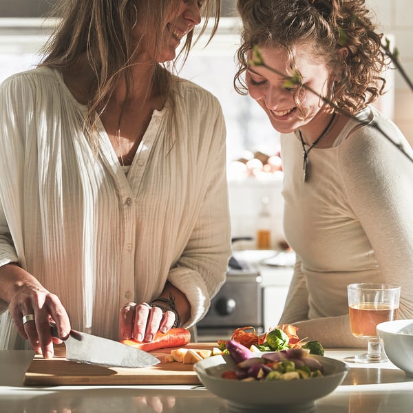 Two people chop vegetables on a cutting board using a knife, with bowls and glasses nearby on a table.