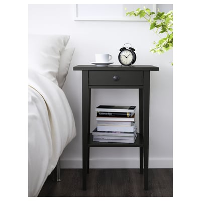Black HEMNES bedside table with books, cup, and alarm clock beside large bed.