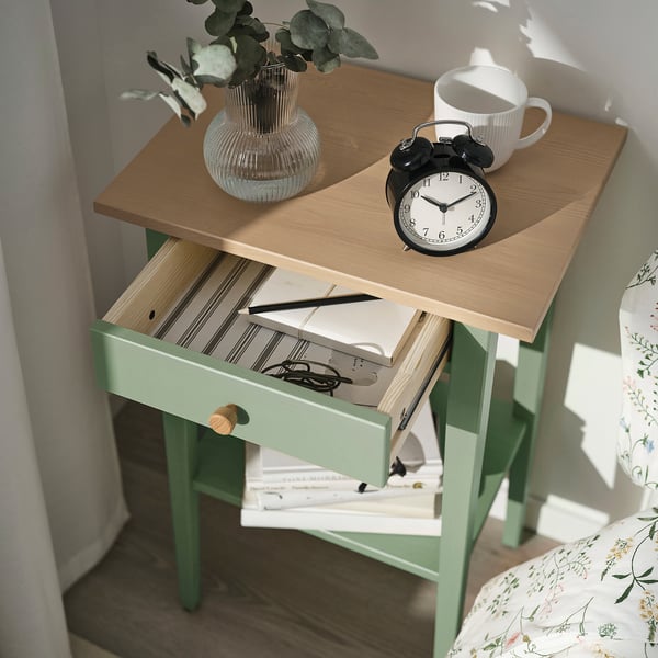 Bedside table with drawer open, showing notebook, pen, and keys. Top has alarm clock and mug. Vase with plant beside.