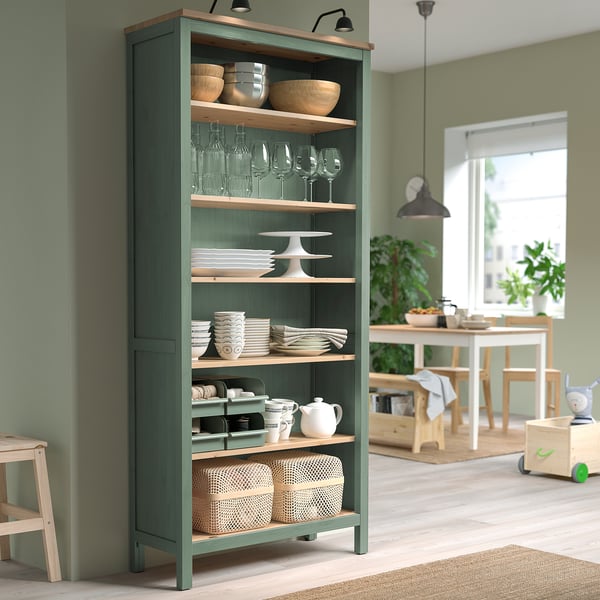 A tall green HEMNES bookcase with wooden shelves. It stands against a green wall, holding various kitchenware and surrounded by wooden furniture.