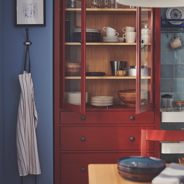A red cabinet with glass doors displays neatly organised dishes and bowls. An apron with blue stripes hangs nearby.