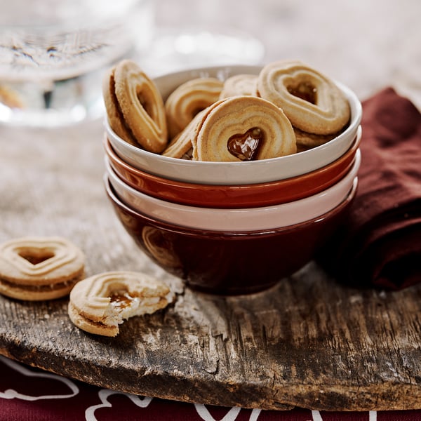 Stacked HÖSTAGILLE stoneware bowls in brown and beige, filled with heart-shaped cookies on a rustic wooden plank.