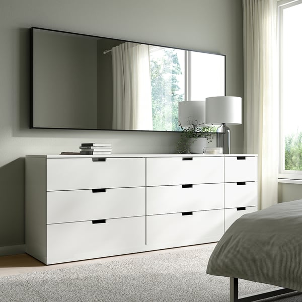 White HOVET mirror above wide dresser in bright bedroom. Mirror reflects greenery, dresser holds books, lamp, and plant.