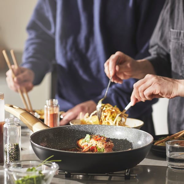Person cooks using black skillet with non-stick surface and wooden spatula. Ingredients and spices surround the cook.