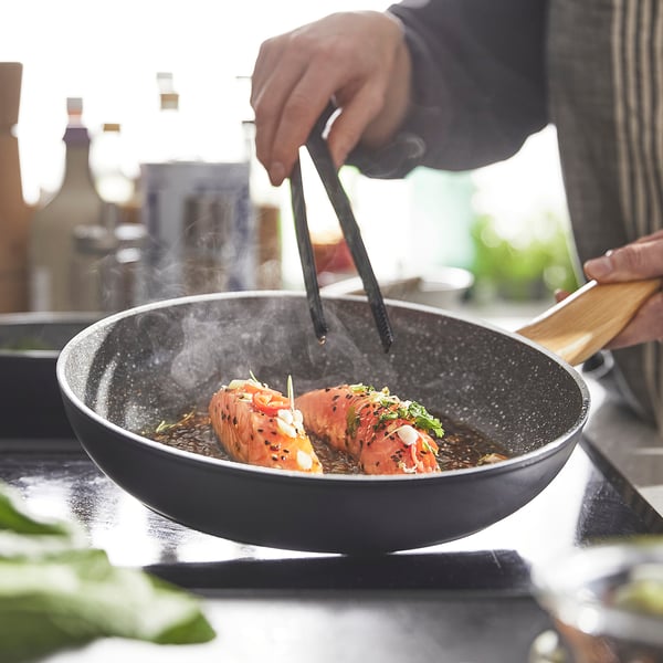 Cook prepares fish in HUSKNUT pan with ceramic non-stick coating, ensuring even heat distribution and easy lifting with grip-friendly handles.