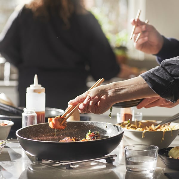Cooking with non-stick pans. One lifts food, another stirs. Bottles and glasses nearby.
