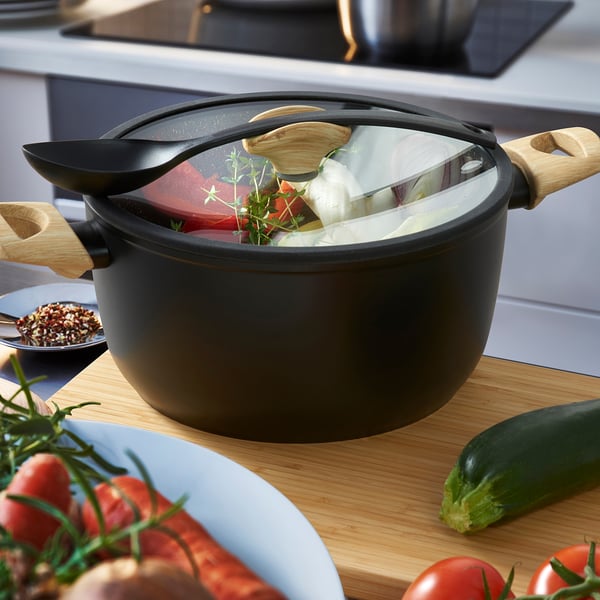 Black saucepan with wooden handles on wooden counter, fresh veggies around, cooking vegetables and herbs inside.