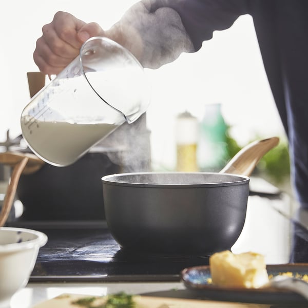 Person pouring liquid from jug into black saucepan on stove. Steam rises, indicating cooking. Wooden utensils nearby.