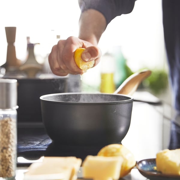 Person squeezes lemon into black saucepan with steam, surrounded by ingredients, likely cooking with heat-resistant, non-stick HUSKNUT cookware.