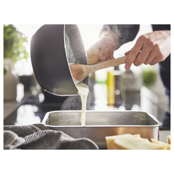 Person pours batter from black pan into metal tray using wooden spatula. Kitchen setting visible.