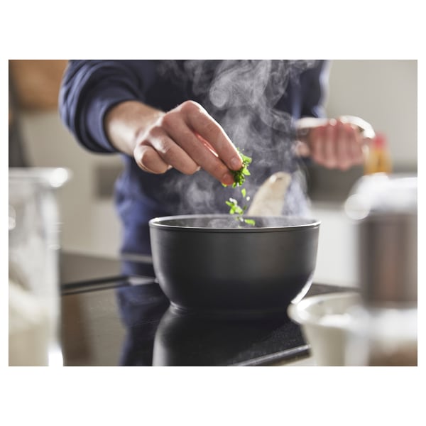 Person seasoning food in a black skillet with steam rising, wearing a blue sweater.