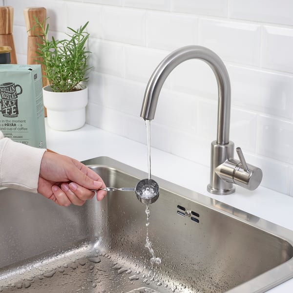 Person fills metal cup under modern chrome kitchen tap. Stainless steel sink, white tiles, and green plant visible.