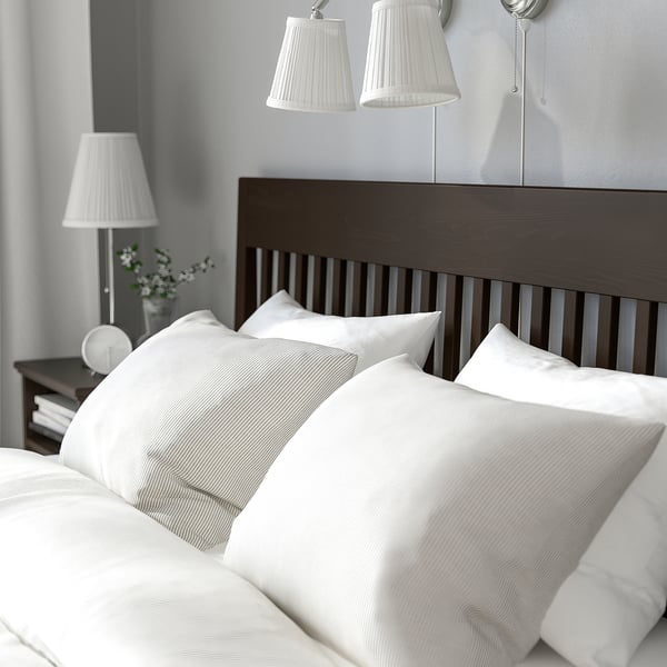 A made bed with dark headboard, white linens, lambswool pillows, comforter, two bedside lamps, and a vase with flowers.