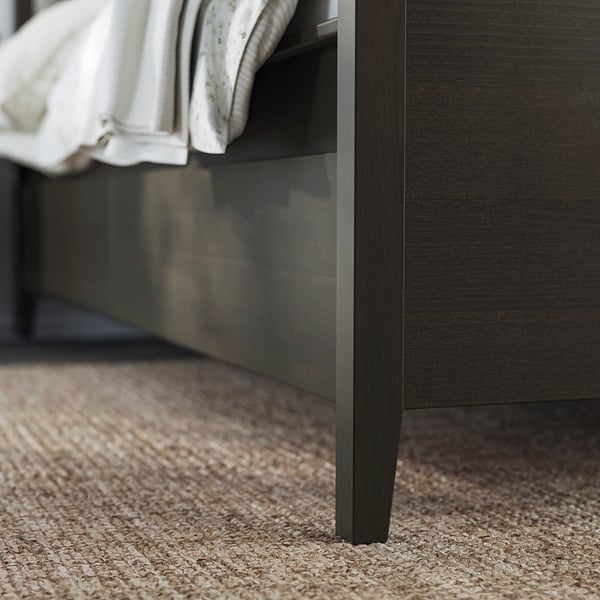 Modern dark wood bed frame with tapered legs on a beige textured rug. Part of the IDANÄS collection.