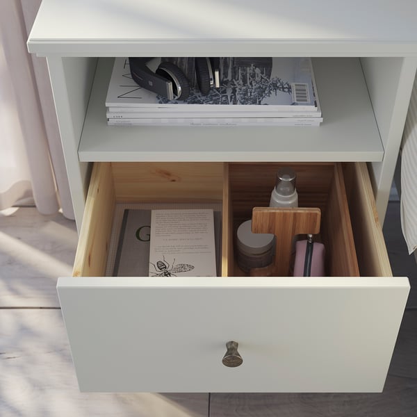 White IDANÄS bedside table with open shelf and drawer storing books and small items.