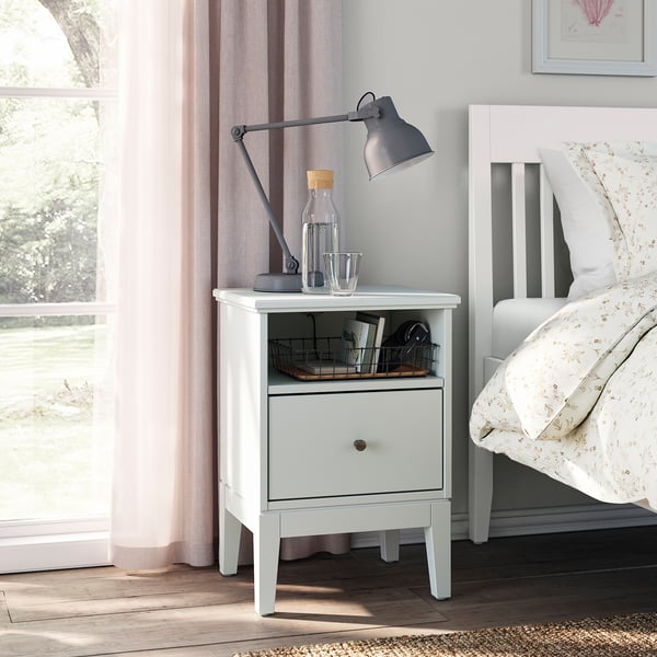 Bedside table named IDANÄS, white, wooden, rectangular with open shelf, drawer, gray lamp, water bottle, glass, and basket.