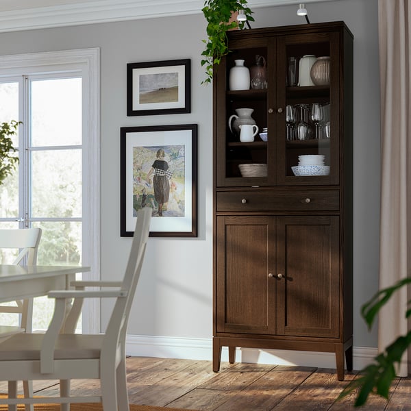 Bright dining room with dark wood IDANÄS cabinet, displaying glassware and vases, next to window with white chairs.