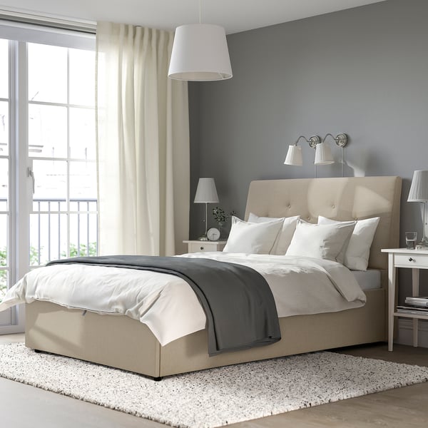 The IDANÄS beige bed with a sloping headboard, white curtains, and wall lamps. The bed is the rooms centerpiece, larger than other items.