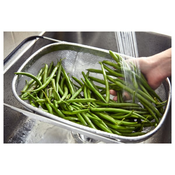 Stainless steel colander with green beans, being rinsed under water in a sink. Adjustable handles visible.