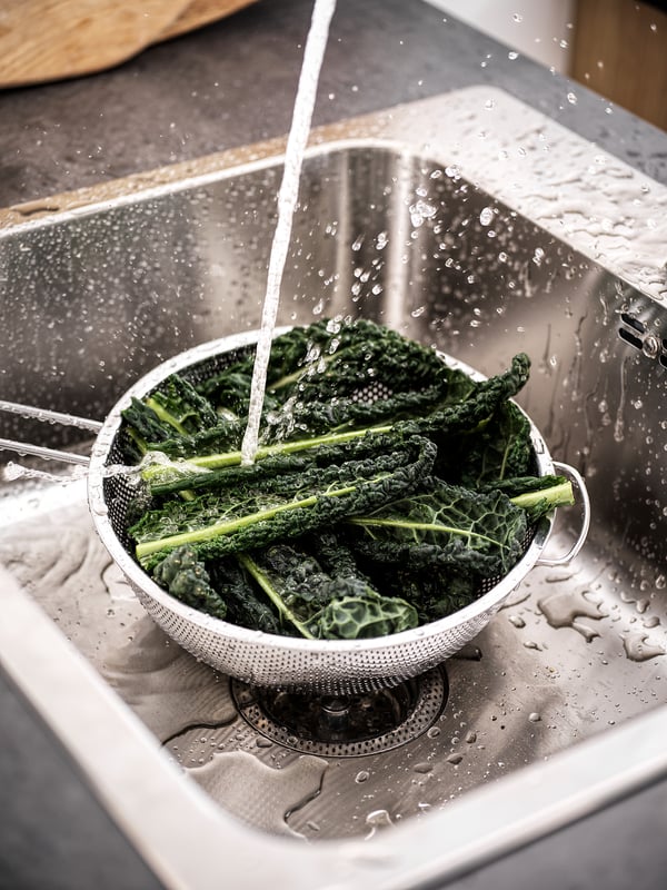 A close-up of green kale being washed in a metal colander over a stainless steel sink. Water streams from above, splashing around the colander.
