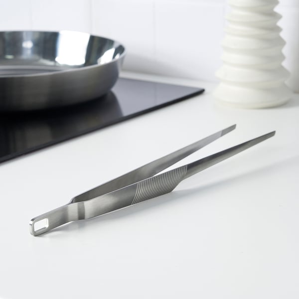 Silver cooking tweezers on a white countertop, with a bowl and a vase nearby. Ideal for gripping and serving food.