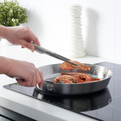 A person is using cooking tweezers to handle shrimp in a pan on a stovetop. The tweezers provide a firm grip for cooking.