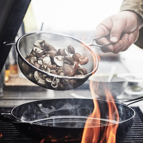A person holds a mesh strainer with mushrooms over a pan with flames.