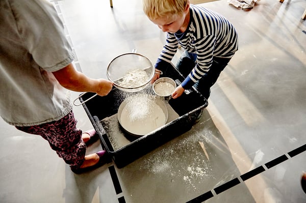 Two people sifting flour into bowl with black strainer.