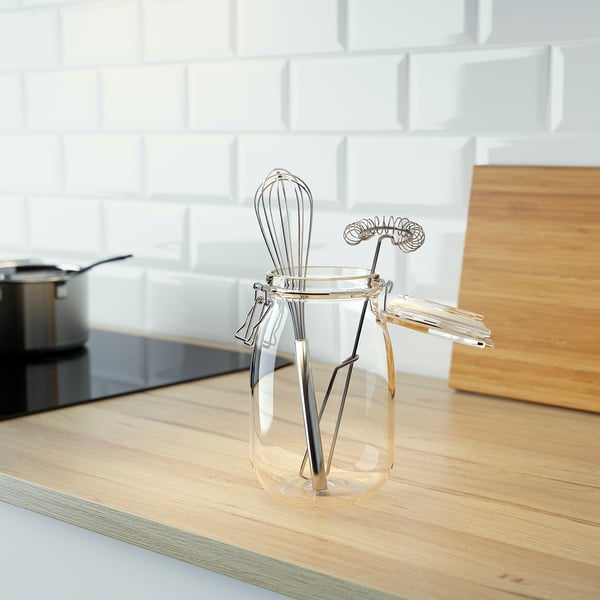 Clear jar holding kitchen utensils: whisk, tongs, and spatula on wooden countertop.