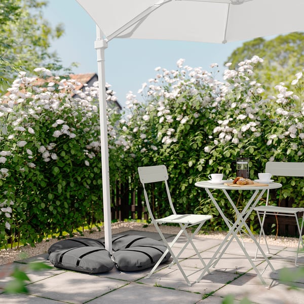Outdoor seating with white folding chairs and table under a white parasol, set against flowering bushes.