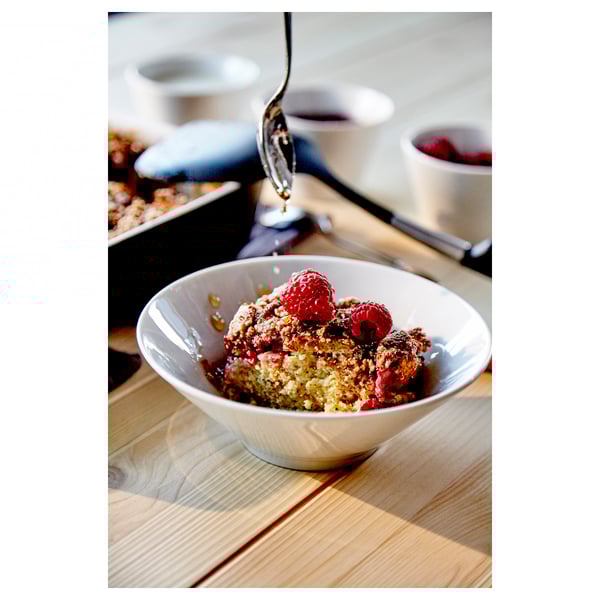 Berry crumble with raspberries in white bowl, wooden table, spoon, coffee.